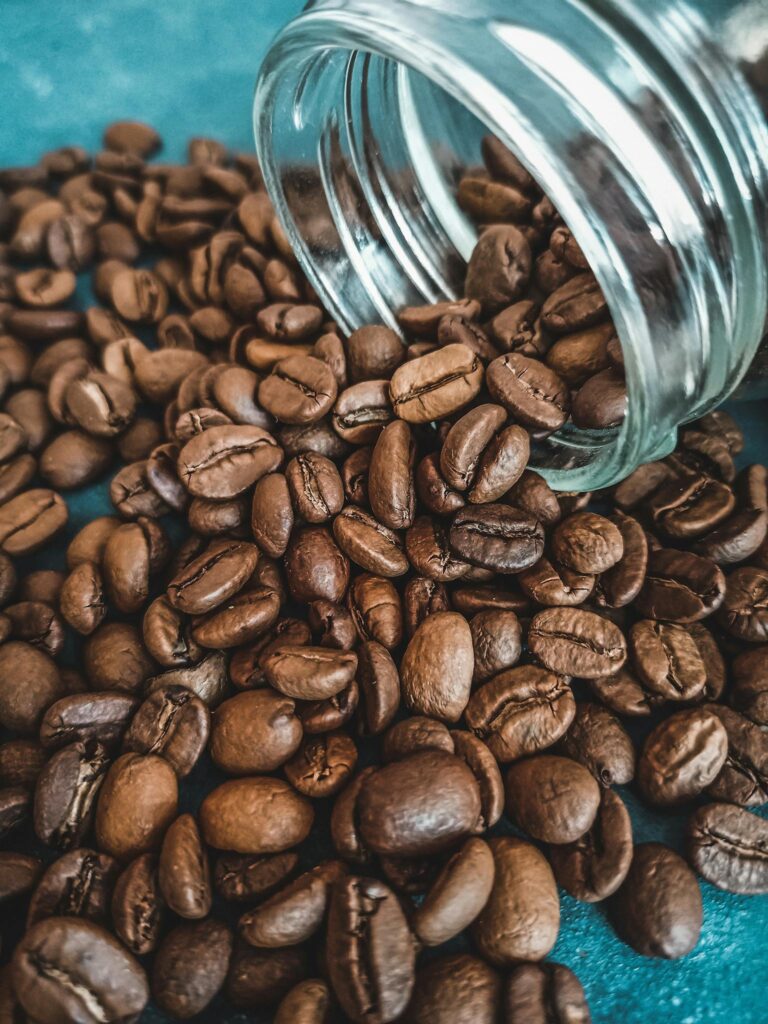 Brown coffee beans spilling from a glass jar onto a blue surface in a close-up shot.